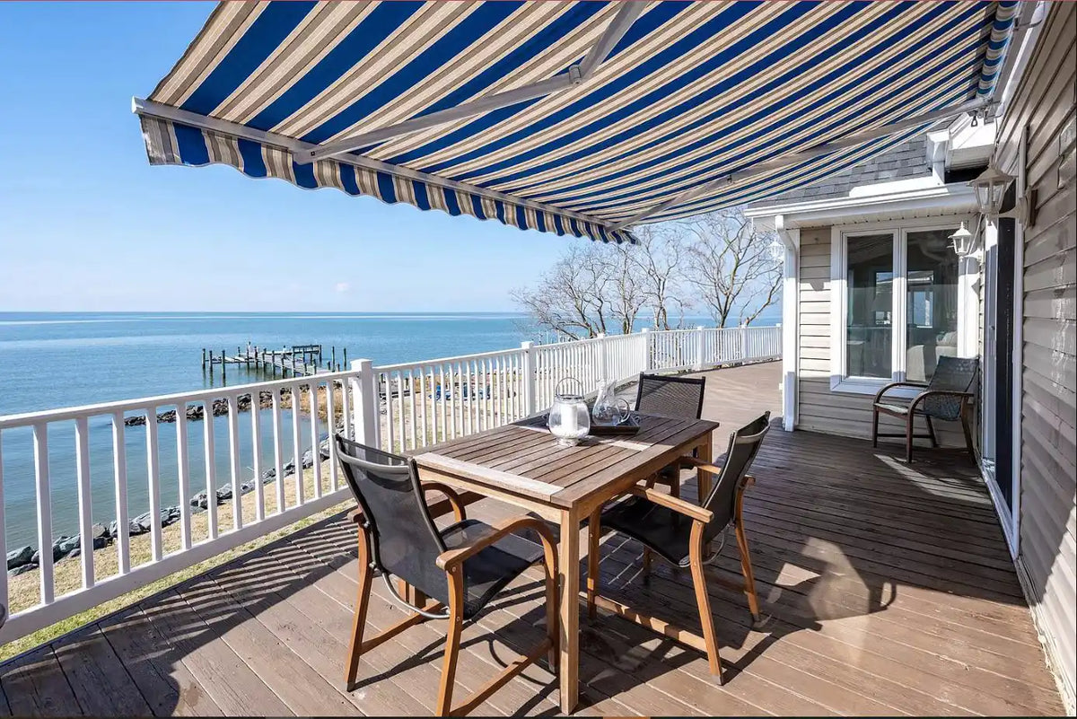 A large blue and cream striped retractable awning covers a wooden deck with a dining table and chairs, overlooking a calm blue ocean and dock.