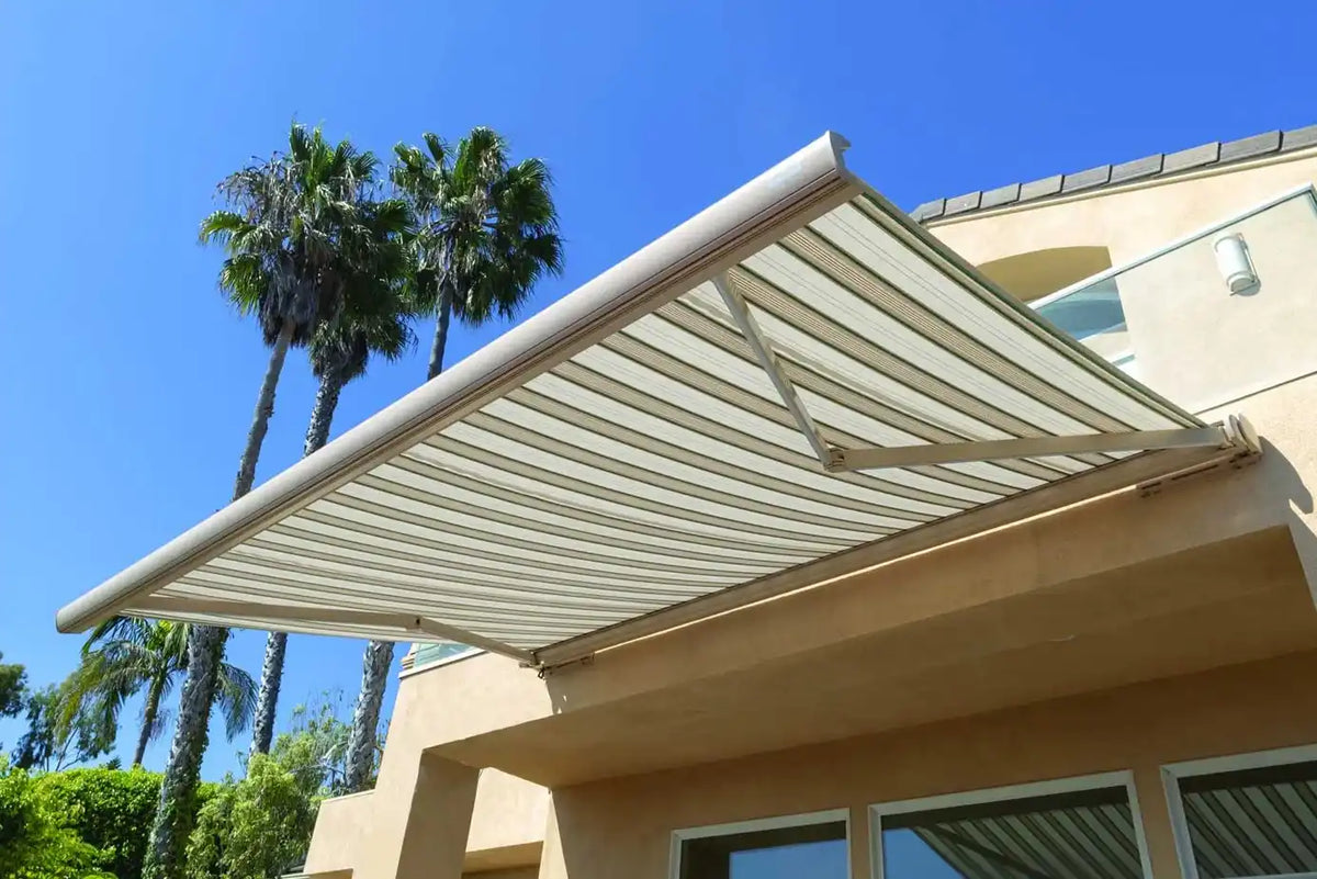A beige striped retractable awning extended over the corner of a stucco house, with palm trees and a clear blue sky in the background.