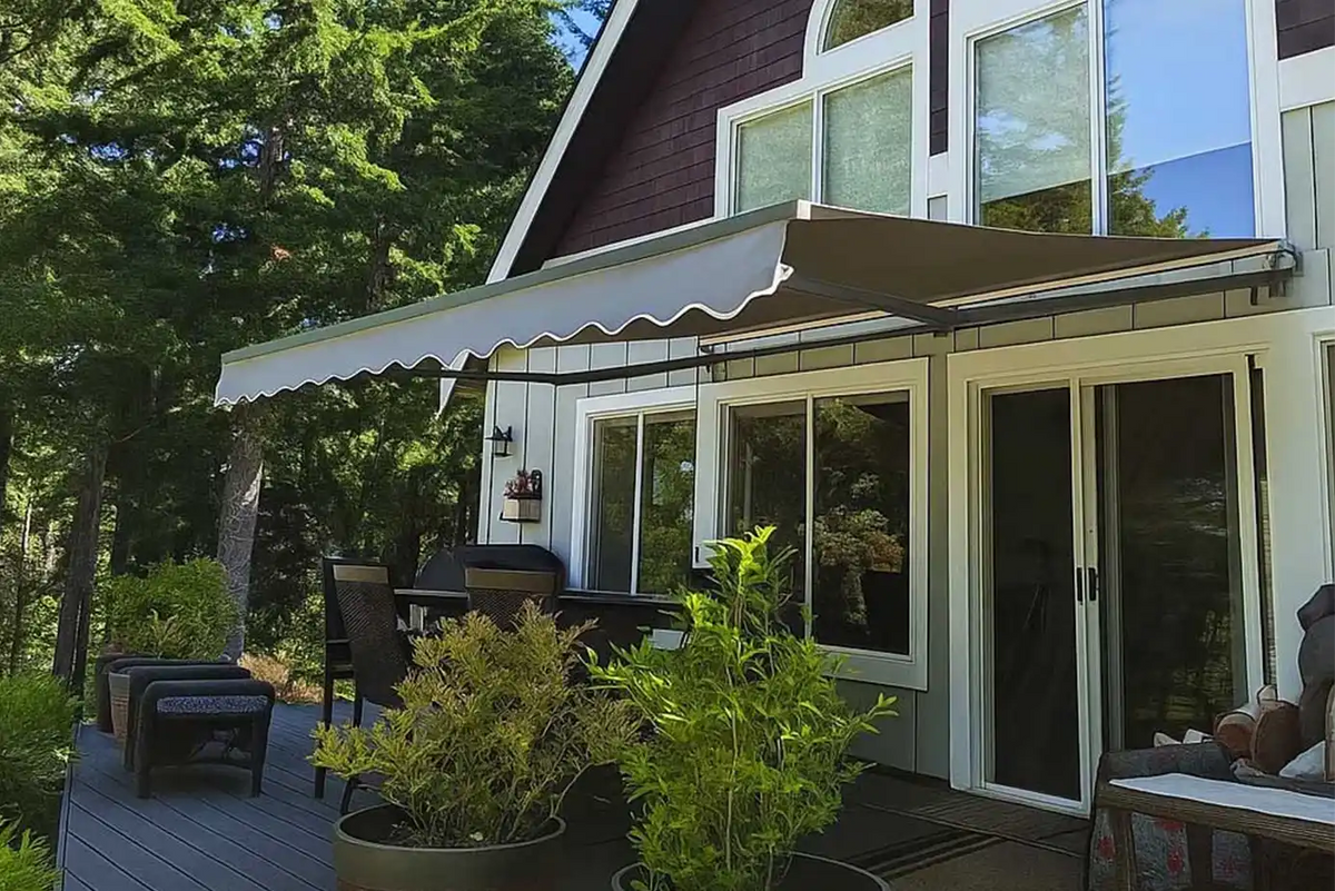 A retractable patio awning with a scalloped white fabric top and dark brown frame, extended over a wooden deck next to a house with dark brown siding and large windows.